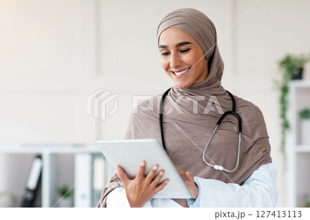 Modern technologies in healthcare industry concept. Smiling muslim woman in hijab doctor with digital tablet posing in her office, copy space. Young lady therapist holding digital pad, clinic interior Modern technologies in healthcare industry concept. Smiling muslim woman in hijab doctor with digital tablet posing in her office, copy space. Young lady therapist holding digital pad, clinic interior 127413113
