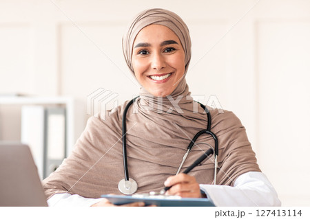 Beautiful muslim woman doctor writing in medical chart and smiling at camera, wearing hijab, sitting at workdesk with modern laptop in her cabinet in clinic or hospital, taking anamnesis, copy space Beautiful muslim woman doctor writing in medical chart and smiling at camera, wearing hijab, sitting at workdesk with modern laptop in her cabinet in clinic or hospital, taking anamnesis, copy space 127413114