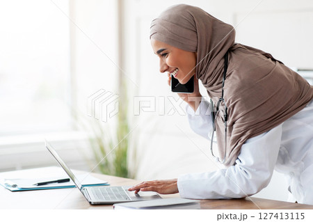 Side view of female doctor young beautiful lady in headscarf arranging appointment on mobile phone, having conversation with patient and looking at her schedule on laptop, clinic interior, copy space 127413115