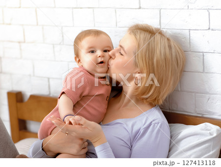 Beautiful mother blonde woman holding and kissing sweet little baby girl, young mom and adorable kid bonding in bed, home interior, copy space. Motherhood, mom's love concept 127413118