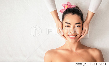 Facial Massage. Attractive young asian lady getting face treatment while relaxing in spa, enjoying beauty procedure, professional beautician touching smiling korean woman's face, top view, copy space Facial Massage. Attractive young asian lady getting face treatment while relaxing in spa, enjoying beauty procedure, professional beautician touching smiling korean woman's face, top view, copy space 127413131