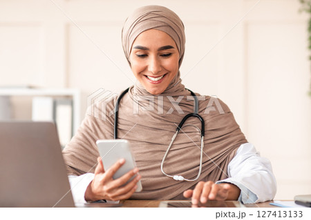 Distance healthcare, telehealth technologies. Happy muslim woman doctor sitting in her cabinet, using digital tablet and mobile phone, copy space. Lady in hijab physician working online from clinic Distance healthcare, telehealth technologies. Happy muslim woman doctor sitting in her cabinet, using digital tablet and mobile phone, copy space. Lady in hijab physician working online from clinic 127413133