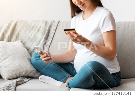 Happy pregnant woman shopping online, using smartphone and credit card at home, young smiling expectant mother purchasing baby clothes from internet while sitting on couch in living room, copy space 127413141
