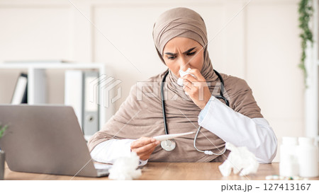 Sick young muslim woman in hijab doctor sneezing nose and looking at thermometer, checking fever, sitting at workdesk with laptop in office at clinic, got sick during COVID-19 pandemic, copy space 127413167