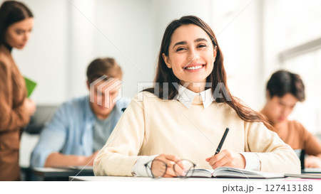 Modern Education Concept. Portrait of smiling female student sitting at desk in classroom at university, taking test or writing notes in her notebook, casual teen looking and posing at camera 127413188