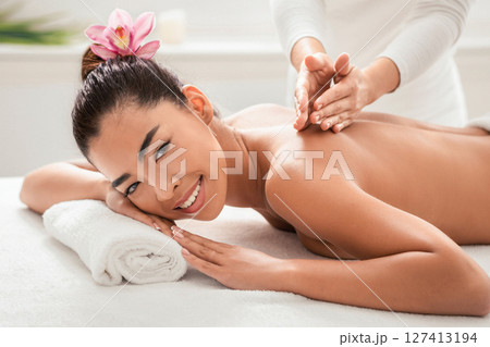 Back Massage. Happy young asian woman getting spa treatment with professional masseur in salon, beautiful korean female with exotic flower in hair relaxing on table and smiling at camera, closeup 127413194