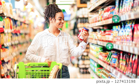 Happy Black Woman Buyer Doing Groceries Shopping Choosing Food And Buying Products Standing With Shop Cart Near Shelf In Modern Supermarket Indoors. Contented Customer In Store Happy Black Woman Buyer Doing Groceries Shopping Choosing Food And Buying Products Standing With Shop Cart Near Shelf In Modern Supermarket Indoors. Contented Customer In Store 127413198