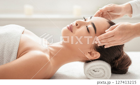Calm young asian lady enjoying relaxing face massage at spa salon, beautiful korean woman lying on massage table and enjoying wellness treatments with professional masseur, side view with copy space 127413215