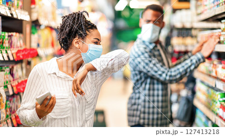 Sick African Woman In Supermarket Coughing In Elbow Wearing Mask Doing Grocery Shopping Buying Food Indoor. Shop Safe, Coronavirus Covid-19 Pandemic Outbreak, Social Distance In Public Places. 127413218