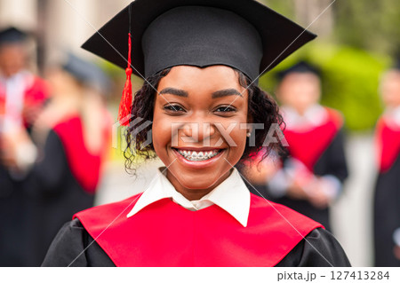 Closeup portrait of happy young black lady student wearing graduating costume. Happy african american female student celebrating graduation with her friends at university campus, blurred background 127413284
