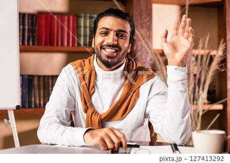 Joyful Indian Teacher Man Waving Hello To Camera Posing Sitting In Classroom With Blackboard Having Online Class Via Video Call. Distant Learning And Teaching, Virtual Lecture And Webinar Concept Joyful Indian Teacher Man Waving Hello To Camera Posing Sitting In Classroom With Blackboard Having Online Class Via Video Call. Distant Learning And Teaching, Virtual Lecture And Webinar Concept 127413292