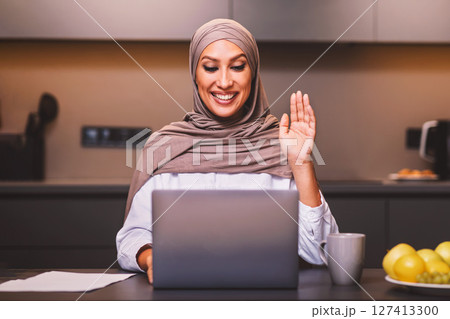 Muslim Lady At Laptop Making Video Call Waving Hello Sitting In Modern Kitchen At Home, Wearing Hijab. Arab Woman Using Computer Communicating Remotely. Female Dietitian Having Online Consultation. Muslim Lady At Laptop Making Video Call Waving Hello Sitting In Modern Kitchen At Home, Wearing Hijab. Arab Woman Using Computer Communicating Remotely. Female Dietitian Having Online Consultation. 127413300