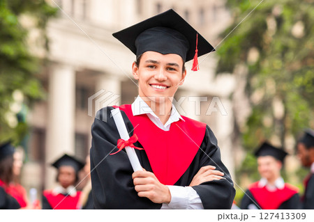 Cheerful guy in graduation costume showing his diploma and smiling at camera, happy male student posing over international group of students at university campus, copy space 127413309