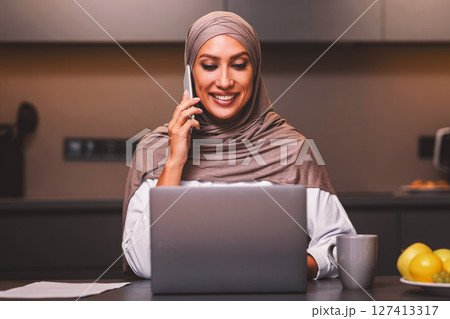 Happy Arab Woman At Laptop Talking On Mobile Phone, Chatting While Browsing Internet Or Working Online Sitting In Kitchen At Home, Wearing Hijab. Successful Muslim Businesswoman Portrait 127413317