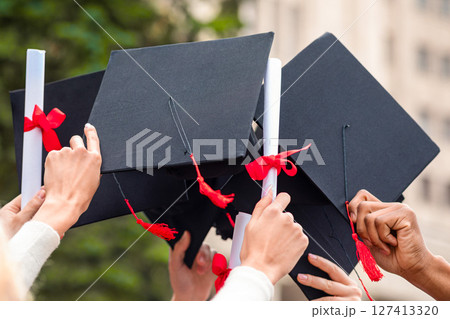 Graduation hats and diplomas raised up in students hands over university or college building, closeup. Cropped of multiracial group of students raising up hands with caps and diploma scrolls 127413320