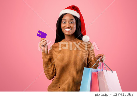 Online Christmas sale concept. Happy black woman in Santa hat holding shopping bags and credit card on pink studio background. Positive African American lady with packed Xmas purchases and bank card Online Christmas sale concept. Happy black woman in Santa hat holding shopping bags and credit card on pink studio background. Positive African American lady with packed Xmas purchases and bank card 127413368