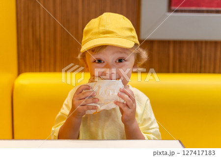 Young child wearing a yellow hat and shirt eats a cheeseburger in a vibrant restaurant, enjoying fast food meal in a casual dining environment. Kids diet, bad food choices, early junk food addiction 127417183
