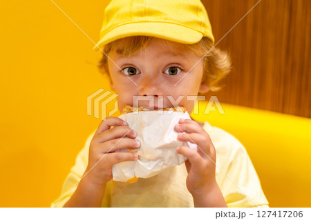 Young child wearing a yellow hat and shirt eats a cheeseburger in a vibrant restaurant, enjoying fast food meal in a casual dining environment. Kids diet, bad food choices, early junk food addiction 127417206
