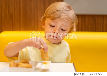 A small child wearing a yellow shirt is eating chicken nuggets in a vibrant environment. Four year old boy enjoying fast food meal. Kids diet, bad food choices, early junk food addiction 127417220
