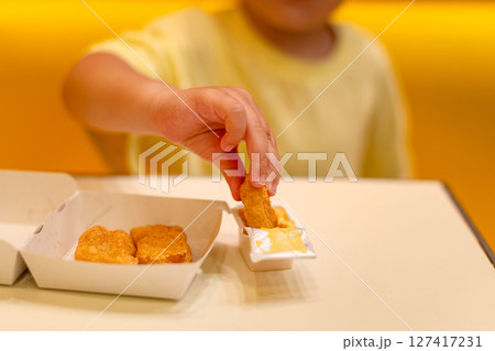 A small child wearing a yellow shirt is eating chicken nuggets in a vibrant environment. Four year old boy enjoying fast food meal. Kids diet, bad food choices, early junk food addiction 127417231