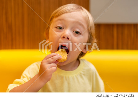 A small child wearing a yellow shirt is eating chicken nuggets in a vibrant environment. Four year old boy enjoying fast food meal. Kids diet, bad food choices, early junk food addiction 127417232