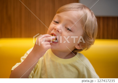 A small child wearing a yellow shirt is eating chicken nuggets in a vibrant environment. Four year old boy enjoying fast food meal. Kids diet, bad food choices, early junk food addiction A small child wearing a yellow shirt is eating chicken nuggets in a vibrant environment. Four year old boy enjoying fast food meal. Kids diet, bad food choices, early junk food addiction 127417235