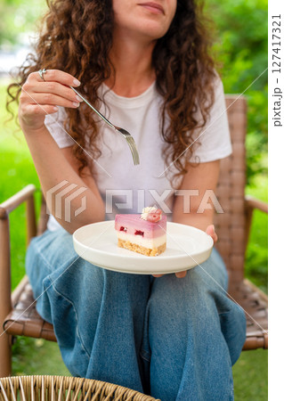 A woman seated at an outdoor table with two plates of desserts, enjoying the scenery and calm surroundings in a peaceful green garden. She smiles thoughtfully, epitomizing comfort and joy in nature. 127417321