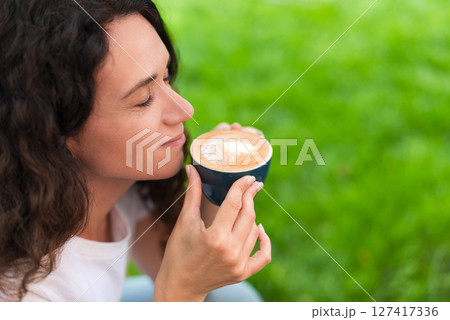A young woman with curly hair holding a cup of coffee, seated outdoors surrounded by vibrant greenery, radiating a calm and introspective mood in a serene and cozy environment. 127417336