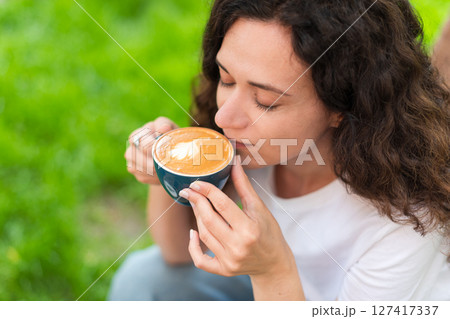 A young woman with curly hair holding a cup of coffee, seated outdoors surrounded by vibrant greenery, radiating a calm and introspective mood in a serene and cozy environment. 127417337