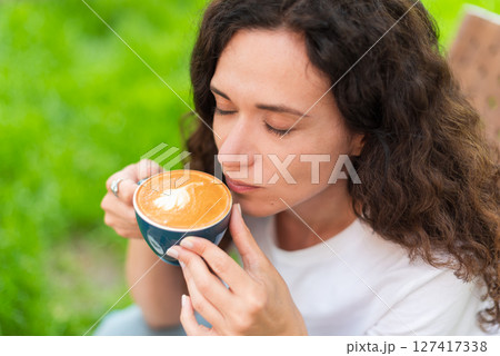 A young woman with curly hair holding a cup of coffee, seated outdoors surrounded by vibrant greenery, radiating a calm and introspective mood in a serene and cozy environment. 127417338