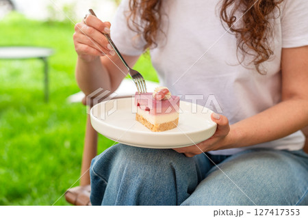 A woman is holding a plate of colorful layered cake while seated in a garden setting. The soft lighting and lush green background enhance the fresh and joyful mood of the scene. 127417353