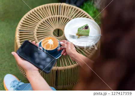 Close-up of a woman holding a cappuccino with latte art and a smartphone during a relaxing outdoors break featuring a plate with a dessert slice on a woven table. 127417359