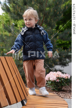 A young child balancing on a curved wooden bench amidst greenery and flowers on a bright day. The scene captures carefree moments in a park. The child is dressed warmly and smiling. A young child balancing on a curved wooden bench amidst greenery and flowers on a bright day. The scene captures carefree moments in a park. The child is dressed warmly and smiling. 127417374