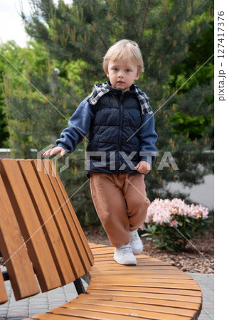 A young child balancing on a curved wooden bench amidst greenery and flowers on a bright day. The scene captures carefree moments in a park. The child is dressed warmly and smiling. 127417376