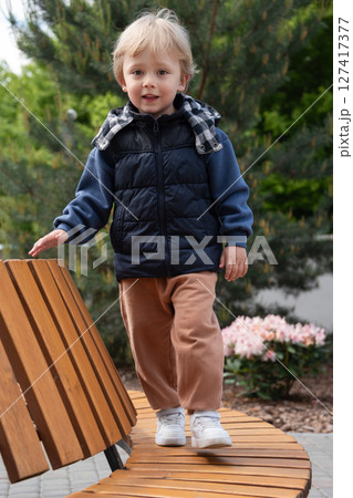 A young child balancing on a curved wooden bench amidst greenery and flowers on a bright day. The scene captures carefree moments in a park. The child is dressed warmly and smiling. A young child balancing on a curved wooden bench amidst greenery and flowers on a bright day. The scene captures carefree moments in a park. The child is dressed warmly and smiling. 127417377