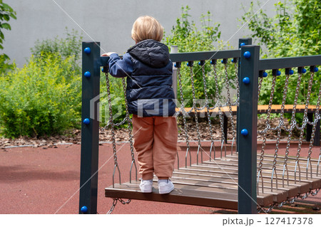 A young child enjoying and exploring a playground activity, walking on a wooden rope bridge in an outdoor park setting, promoting childhood fun, motor skills, and healthy outdoor recreation. A young child enjoying and exploring a playground activity, walking on a wooden rope bridge in an outdoor park setting, promoting childhood fun, motor skills, and healthy outdoor recreation. 127417378