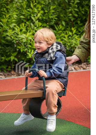 Child enjoying a seesaw ride in a sunny outdoor playground area. The setting includes nature surrounding a colorful playground, depicting joy and bonding during leisure time. 127417390