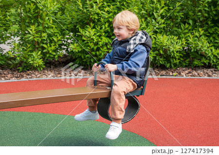 Child enjoying a seesaw ride in a sunny outdoor playground area. The setting includes nature surrounding a colorful playground, depicting joy and bonding during leisure time. 127417394