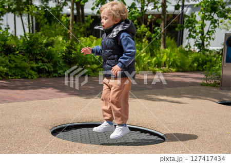 A young child jumping on a trampoline at an outdoor park surrounded by greenery. The child shows excitement and joy while playing, embodying youthful energy and fun in the sunlit environment. A young child jumping on a trampoline at an outdoor park surrounded by greenery. The child shows excitement and joy while playing, embodying youthful energy and fun in the sunlit environment. 127417434