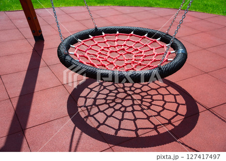 A red net swing casts an intricate shadow on playground tiles, creating visual interest with vibrant colors and geometric shapes in a sunny outdoor park setting. 127417497