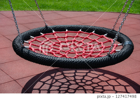 A red net swing casts an intricate shadow on playground tiles, creating visual interest with vibrant colors and geometric shapes in a sunny outdoor park setting. 127417498