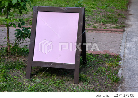 A blank pink chalkboard standing on a sidewalk outdoors, ready for business messages. The board is surrounded by greenery, making it ideal for attracting the attention of pedestrians passing by. 127417509