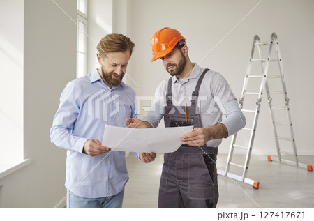 Architect in uniform and hardhat, holding house construction blueprint, talking with man client Architect in uniform and hardhat, holding house construction blueprint, talking with man client 127417671