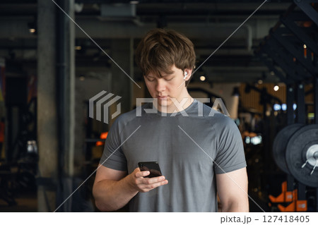 Young caucasian sporty man relax and standing with mobile phone, listen music headphones and texting at gym. Bodybuilder during training. Fitness healthy lifestyle concept. 127418405