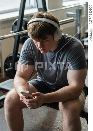 Young caucasian sporty man relax and sitting with mobile phone, listen music headphones and texting at gym. Bodybuilder during training. Fitness healthy lifestyle concept. Vertical format. Young caucasian sporty man relax and sitting with mobile phone, listen music headphones and texting at gym. Bodybuilder during training. Fitness healthy lifestyle concept. Vertical format. 127418406