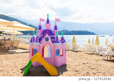 Pink plastic fairy-tale castle on beach sand. Seaside playground with loungers, umbrellas, and mountains in background. Pink plastic fairy-tale castle on beach sand. Seaside playground with loungers, umbrellas, and mountains in background. 127418586