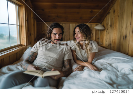 A man and a woman are lying on a bed, with the man reading a book 127420034