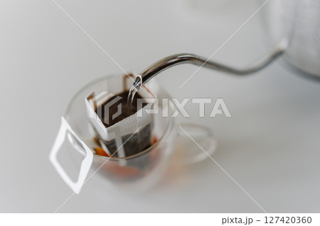 Close-up of hot water pouring from kettle into drip coffee bag placed in glass cup, offering simple and convenient way to brew fresh cup of coffee. Concept of quick way to enjoy fresh cup of coffee. Close-up of hot water pouring from kettle into drip coffee bag placed in glass cup, offering simple and convenient way to brew fresh cup of coffee. Concept of quick way to enjoy fresh cup of coffee. 127420360