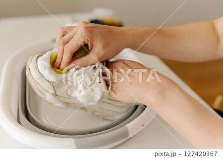 Male carefully cleaning dirty white sneakers with yellow sponge and soap foam in small foldable basin, taking care of footwear hygiene and cleanliness at home in bathroom, close-up. Male carefully cleaning dirty white sneakers with yellow sponge and soap foam in small foldable basin, taking care of footwear hygiene and cleanliness at home in bathroom, close-up. 127420367