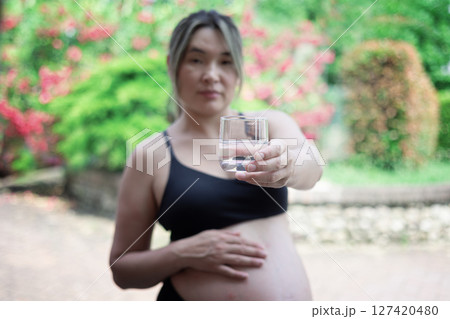 Pregnant woman holding a glass of fresh water, promoting a healthy lifestyle for both herself and her baby during pregnancy, standing in a serene garden setting 127420480